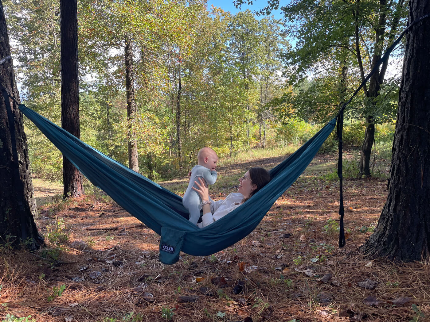 A woman holds a baby while in an ENO TravelNest Hammock. They are in the woods.