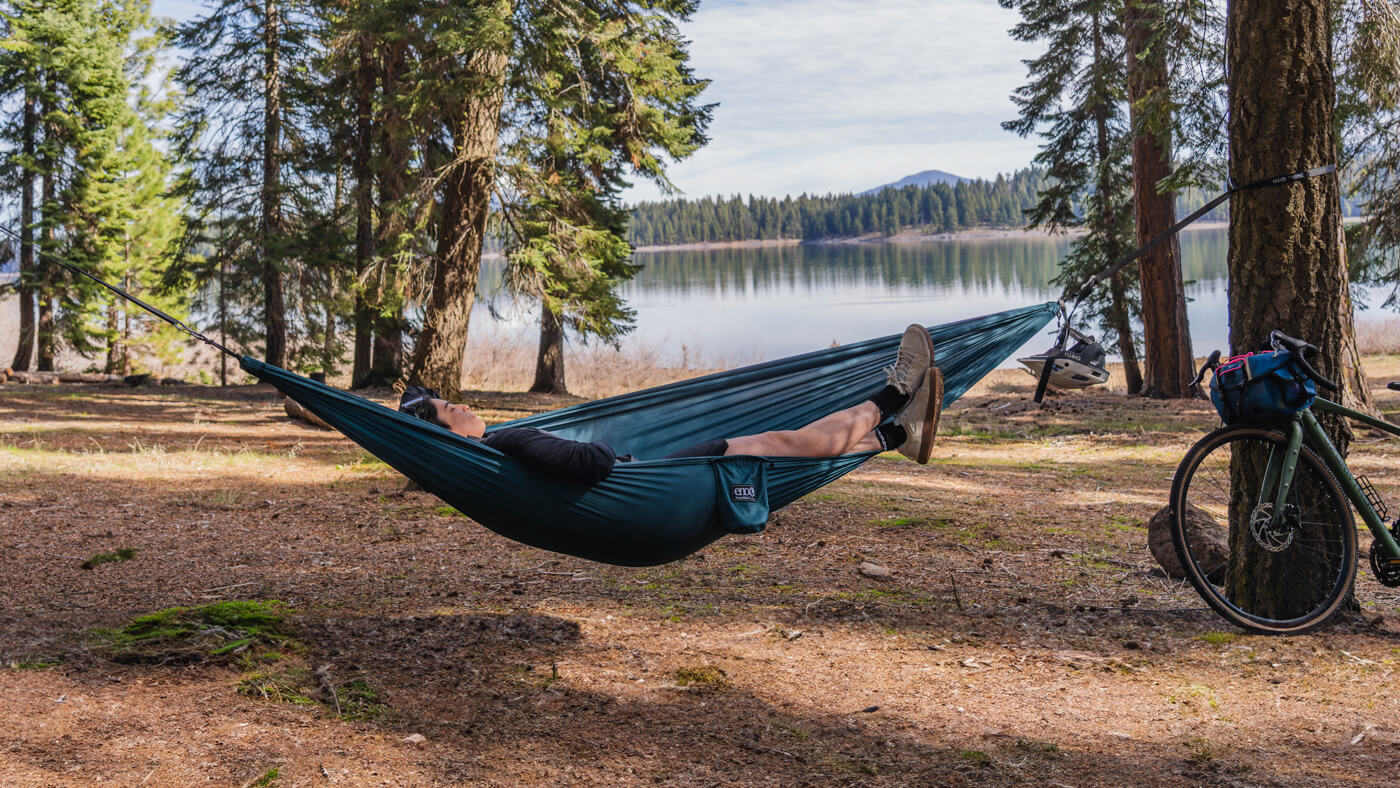 A woman lays in her ENO TravelNest Hammock near a lake with her bike resting on a nearby tree