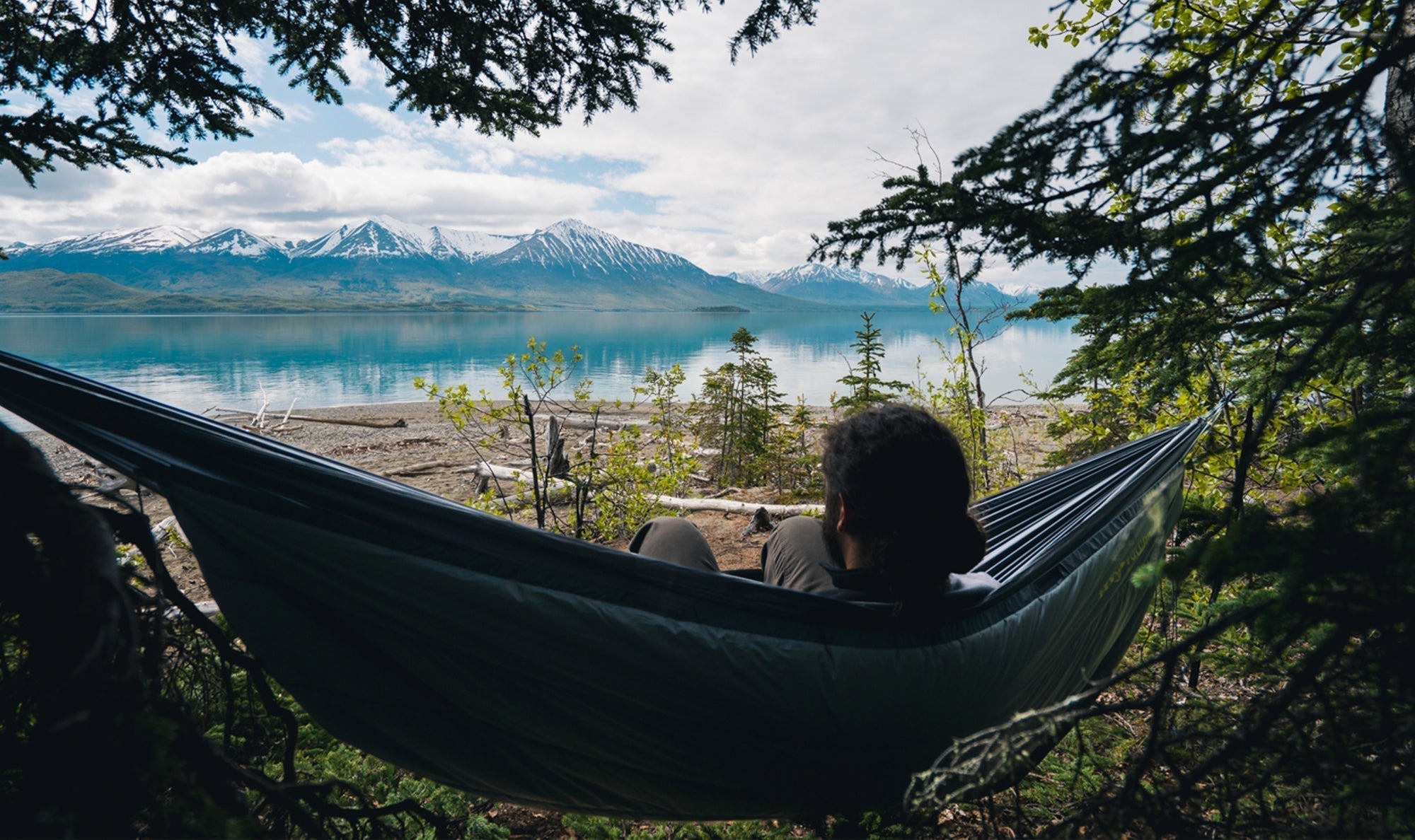 A man looks out at a lake and snowy mountain range while sitting in his ENO hammock