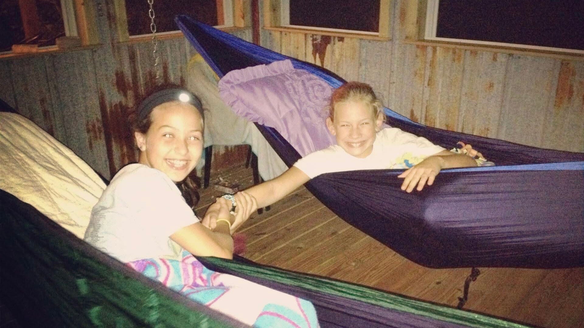 Two girls sitting in hammocks hold hands as they smile for their photo to be taken