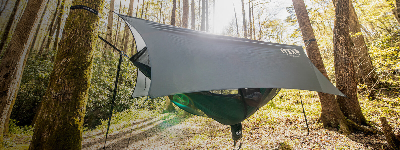 A person laying in an ENO hammock tent consisting of hammock, rain tarp, and bug net set up between two trees in a forest.