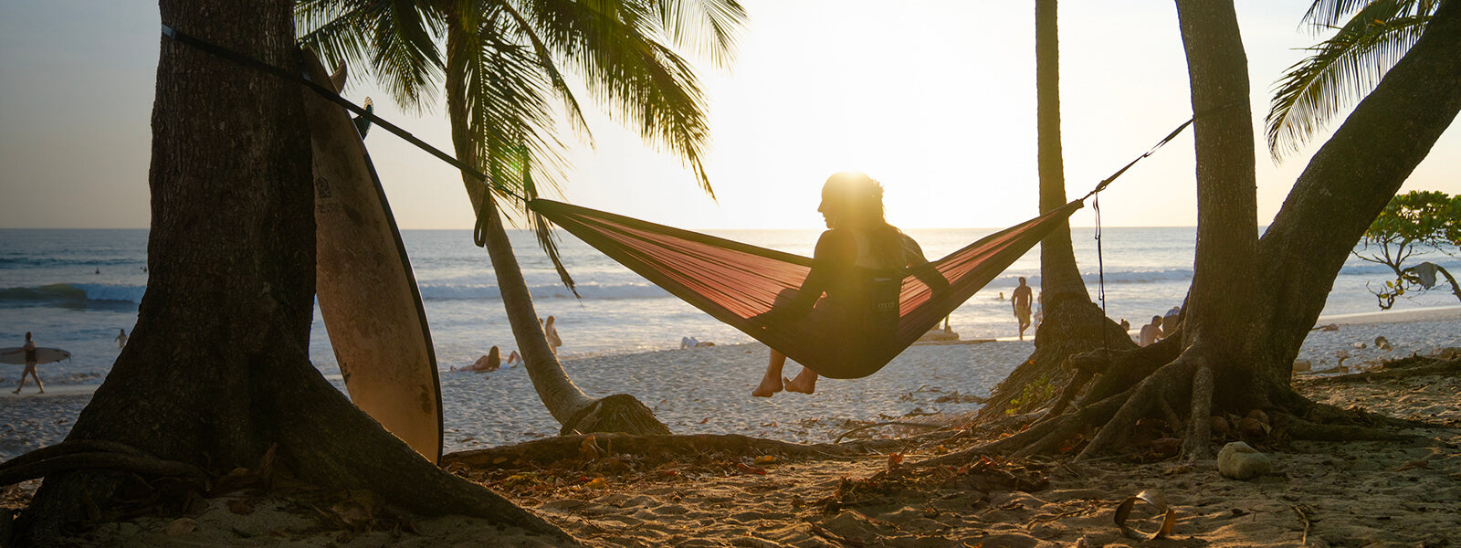 A person sitting in an ENO portable hammock set up between two trees with beach sunset in the background.