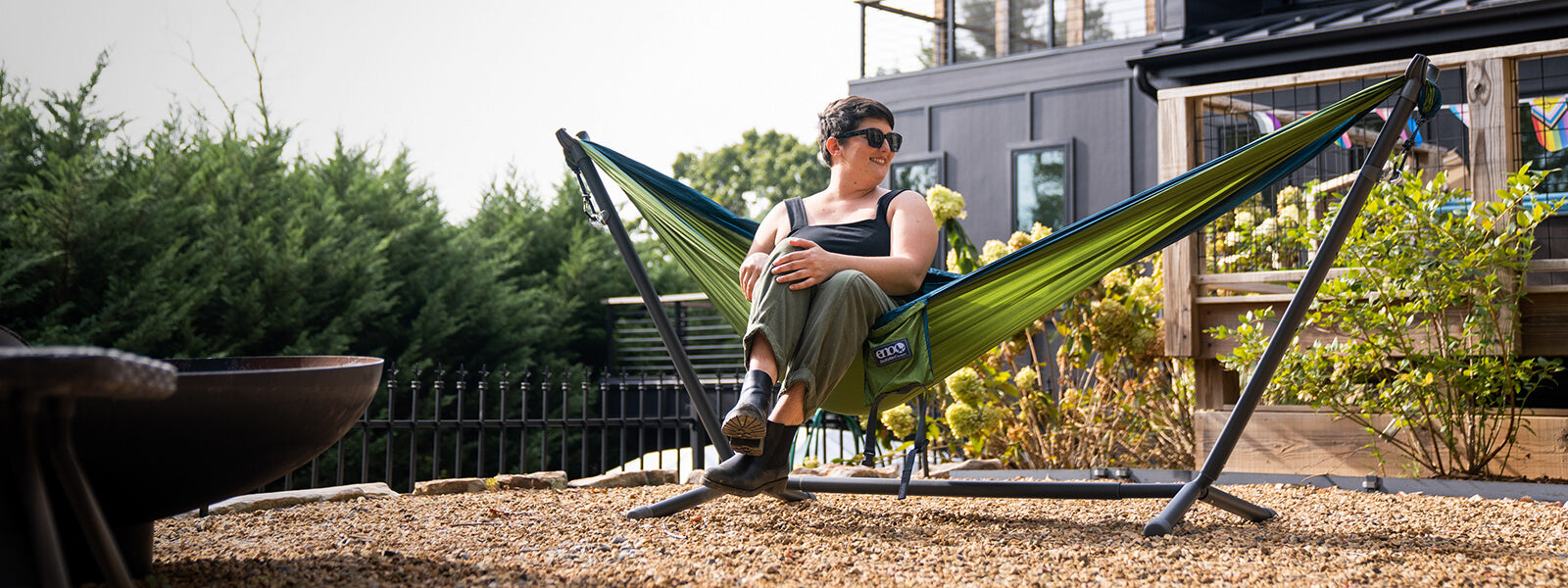 A person sitting in a backyard hammock set up on the ENO Parkway Adjustable Hammock Stand in front of a house.