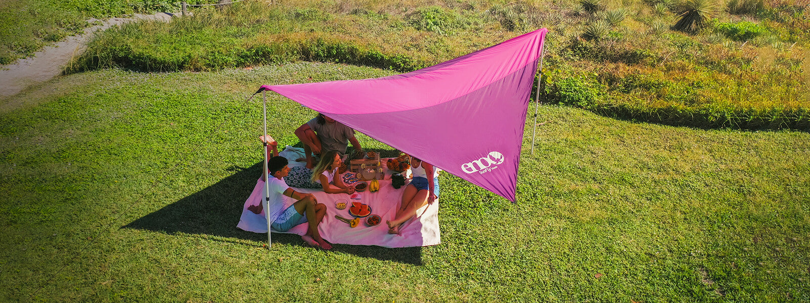 A group of friends sit under a ENO SunFly Shade setup in the park for a picnic.