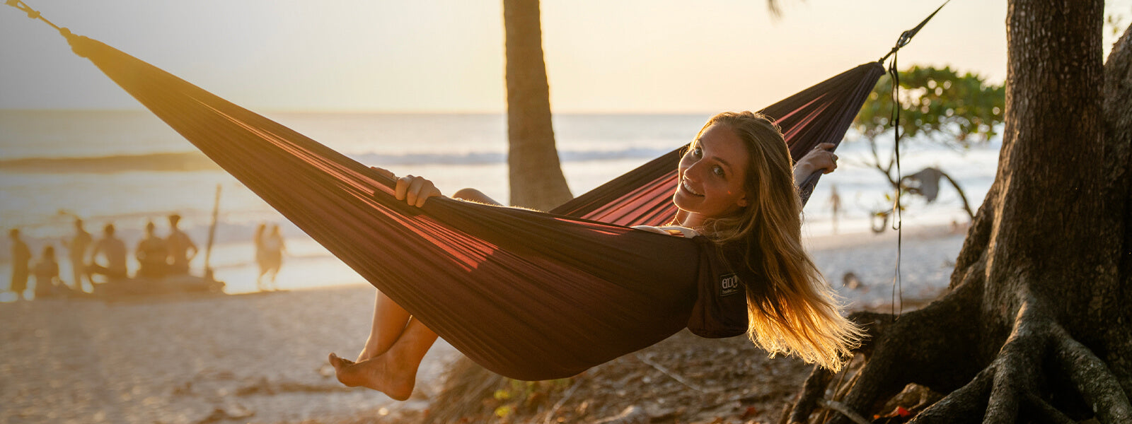 A women looks back at camera over her shoulder on beach in an ENO portable hammock set up between two trees.   