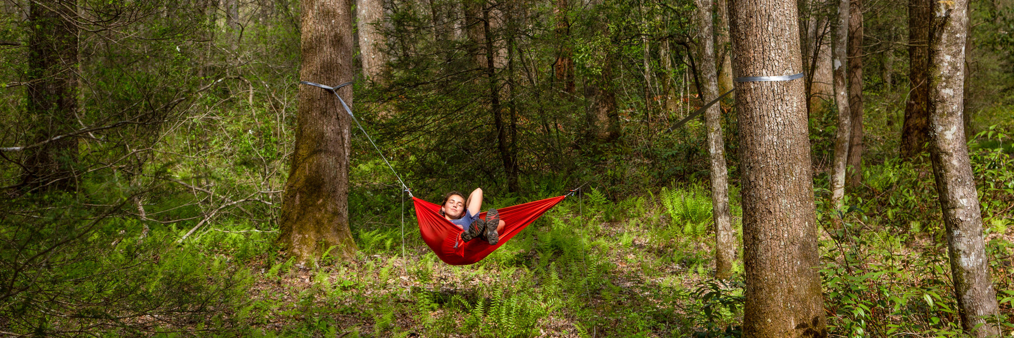 A person laying back in an ENO ultralight hammock relaxing between two trees in a forest.