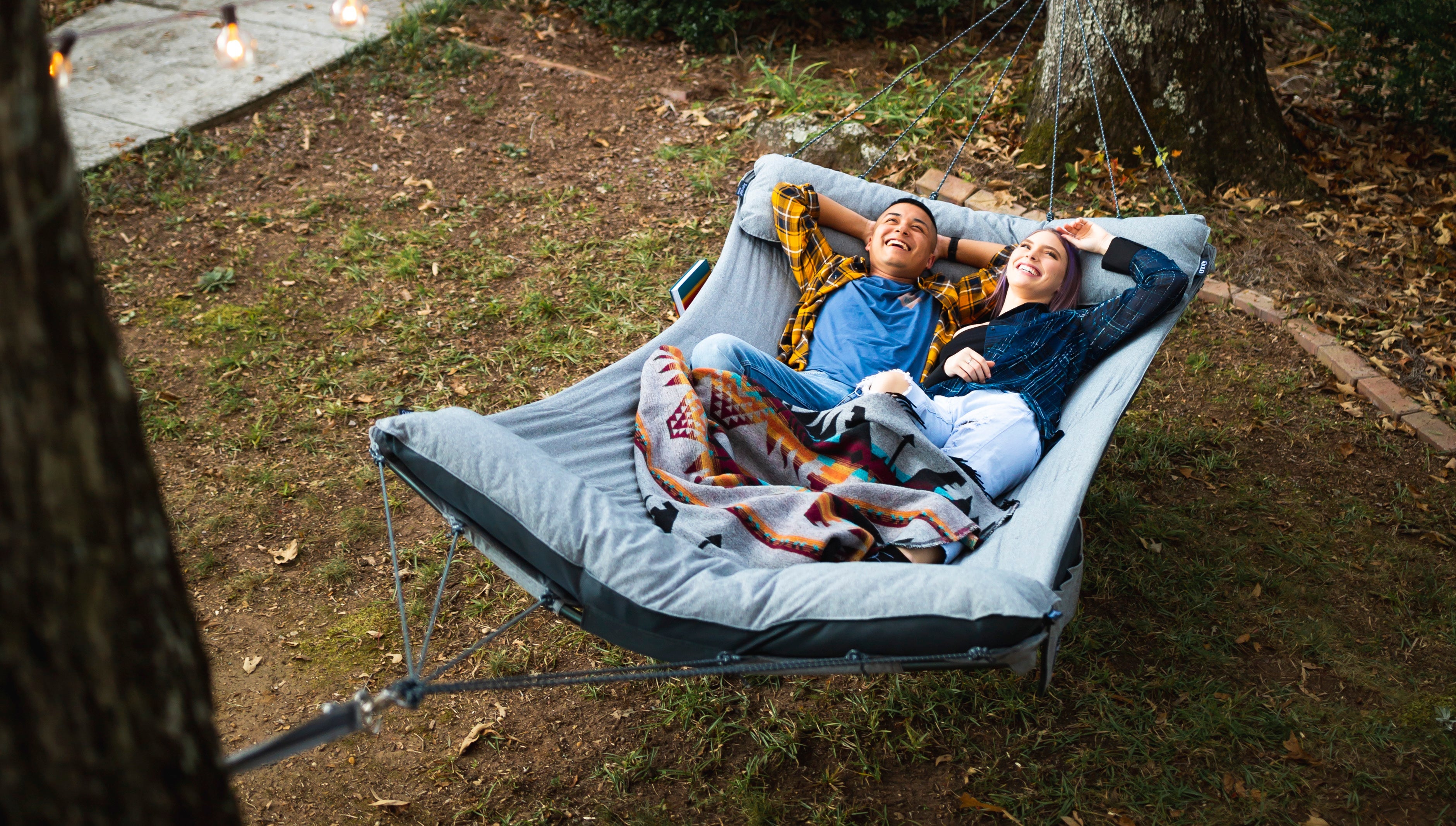 A couple lie smiling looking up in an ENO backyard hammock set up between two trees.