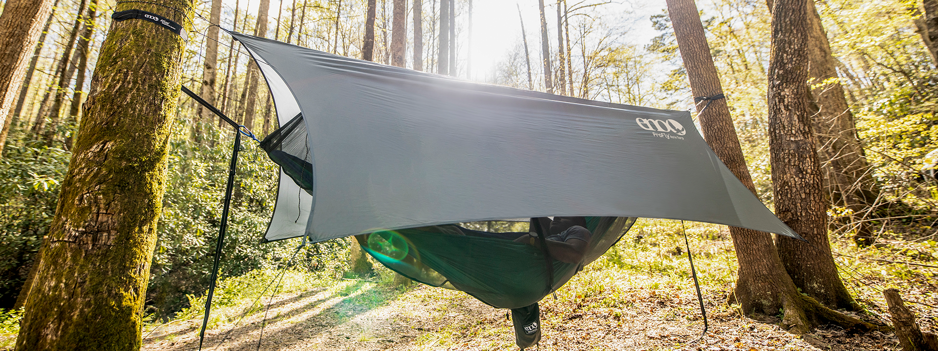 A person laying back in an ENO hammock tent consisting of hammock, rain tarp, and bug net set up between two trees in forest.