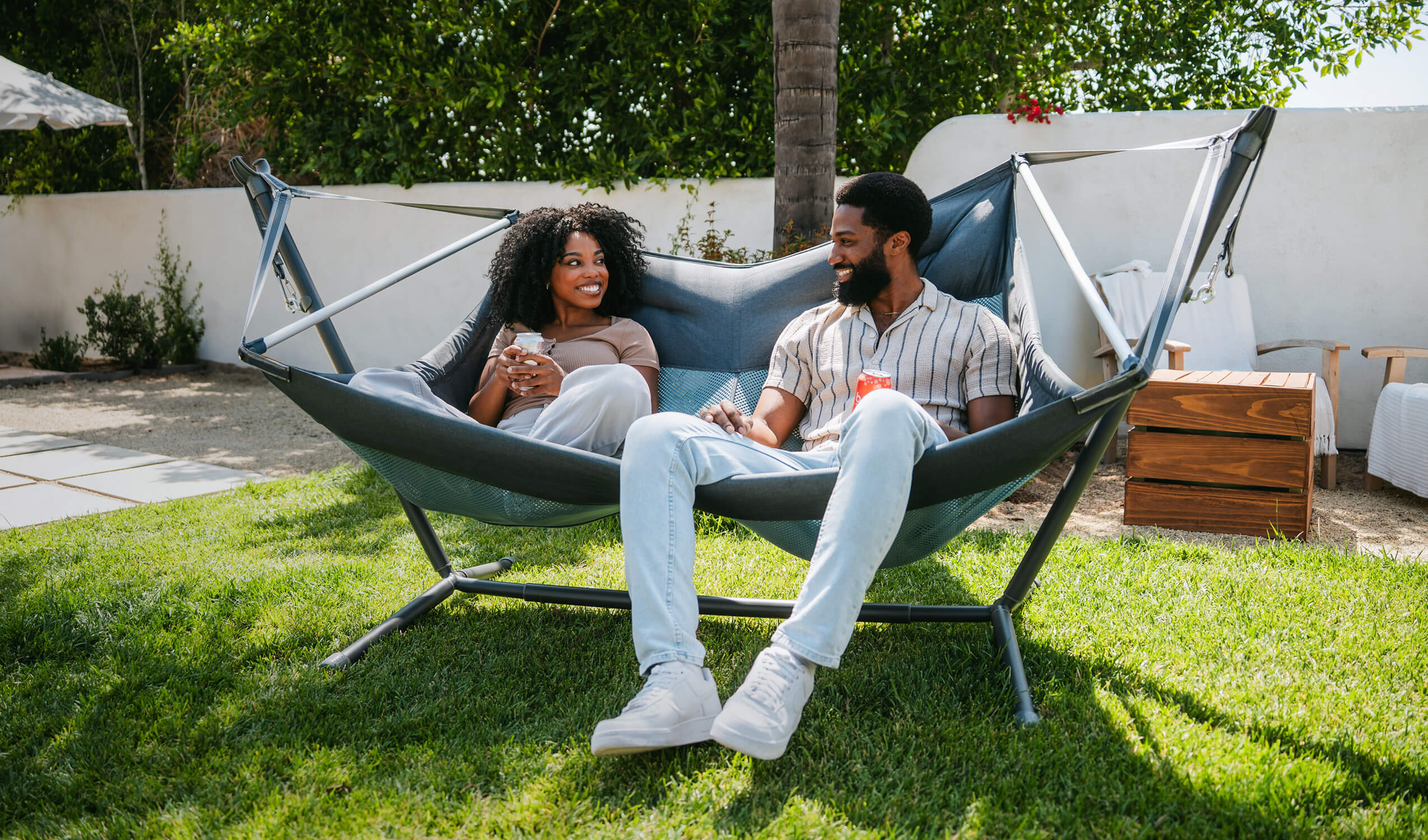 A couple in a backyard environment sitting, smiling at each other while sitting in the ENO DoubleLoft Hammock Chair connected to the Parklite Adjustable Hammock Stand.  