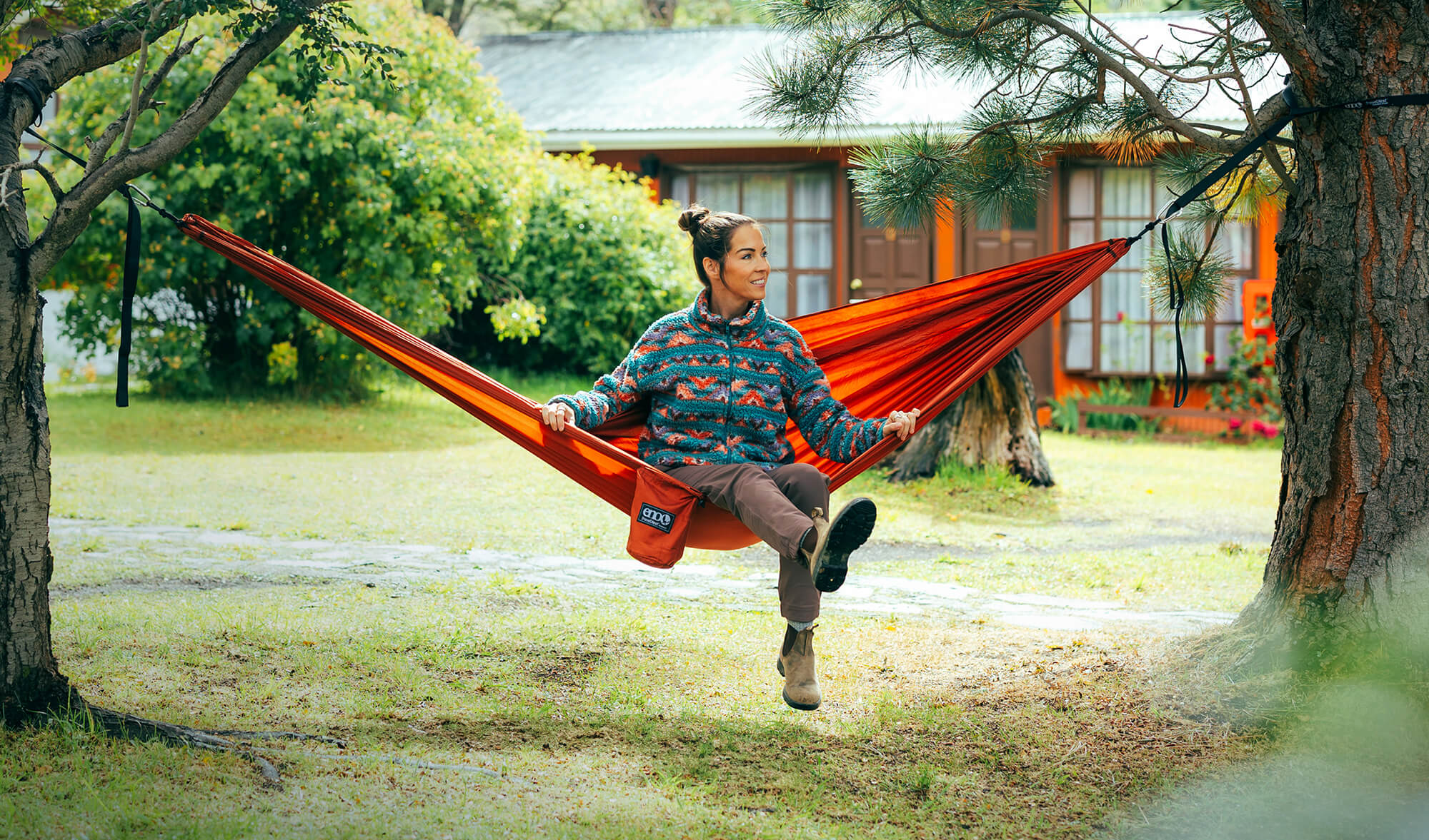 Person sitting in a Canyon TravelNest Hammock + Staps combo in a park-like setting with a house in the background.