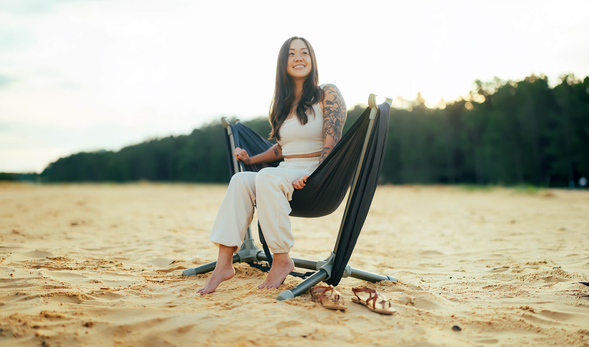 Woman sitting in a hammock in the Parklite Hammock Chair Stand in Agave on a sandy beach with trees in the background