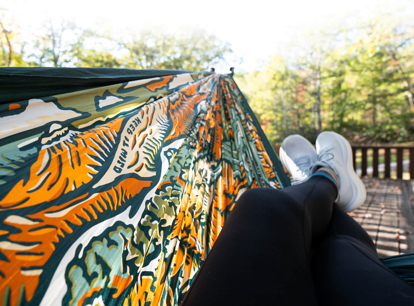 Person laying in a colorful ENO | American Hiking Society Giving Back Hammock with their legs handing out on a porch setting