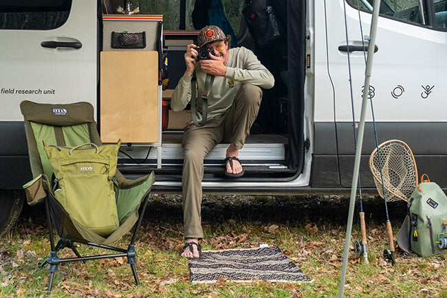 A person sits on the floor of their van while taking a photo on a camera while belonging dress the exterior of the van.