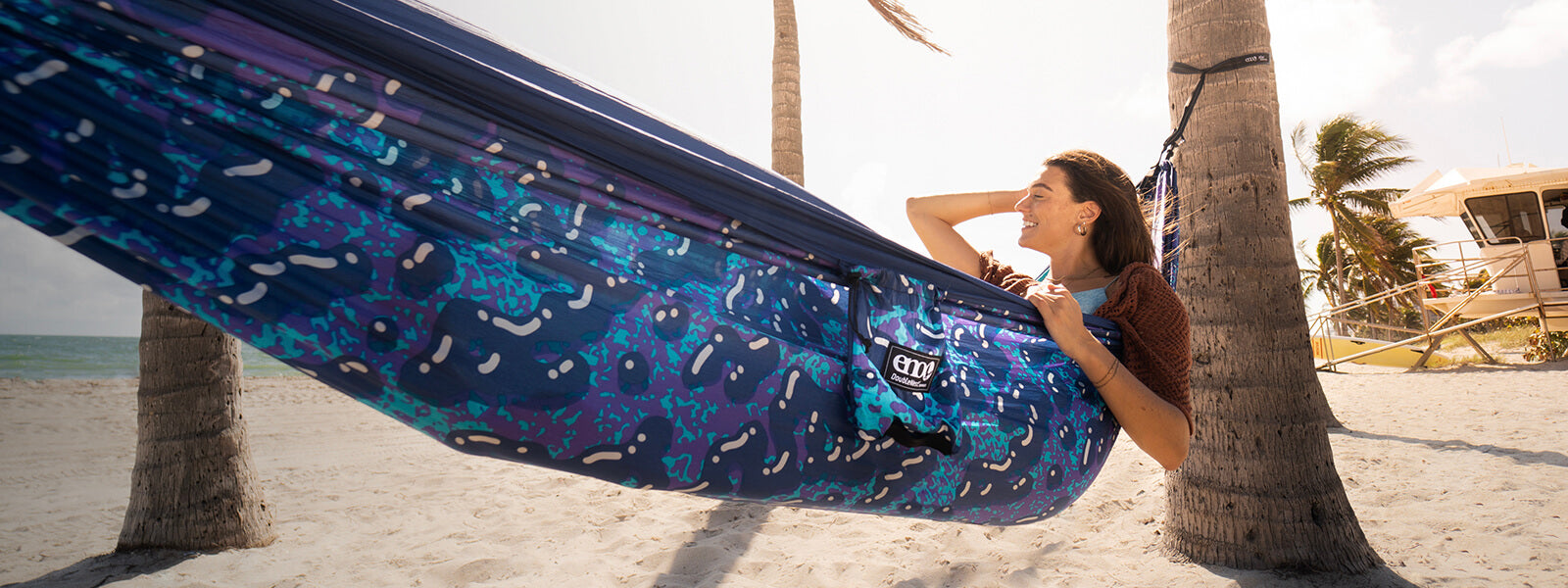 Women sitting up in ENO DoubleNest Hammock Print, smiling as they run their hand through their hair with sand and beach in the background.