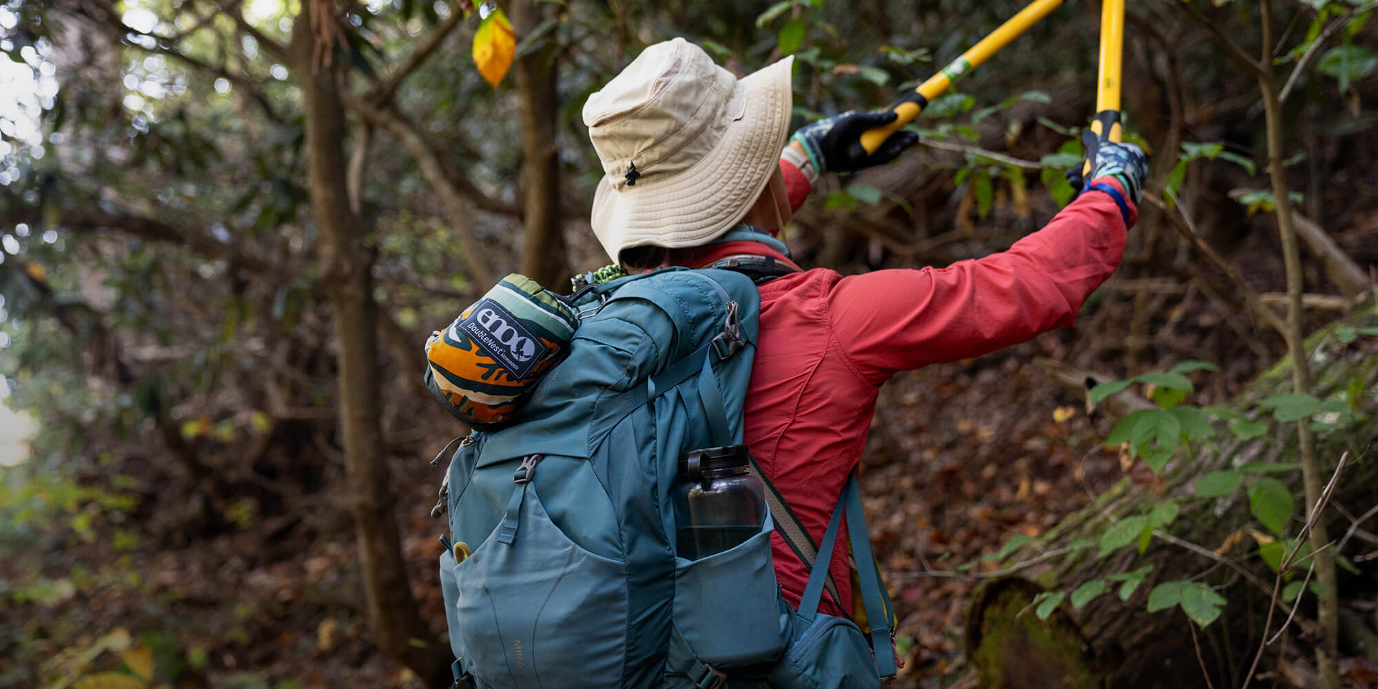 A person trims the forest trail while wearing a backpack with a packed ENO Hammock attached. 