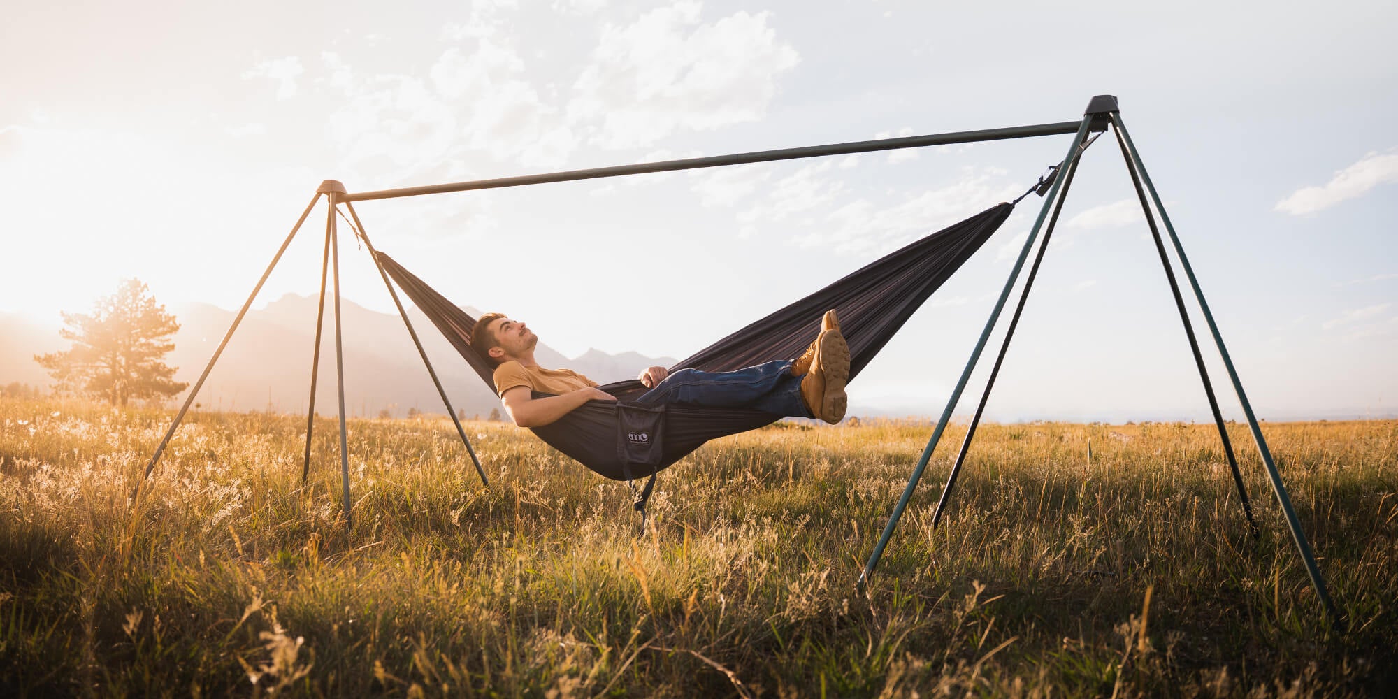 Young man laying back in ENO DoubleNest Hammock attached to the Nomad Hammock Stand under a golden sunset in large grassy field.