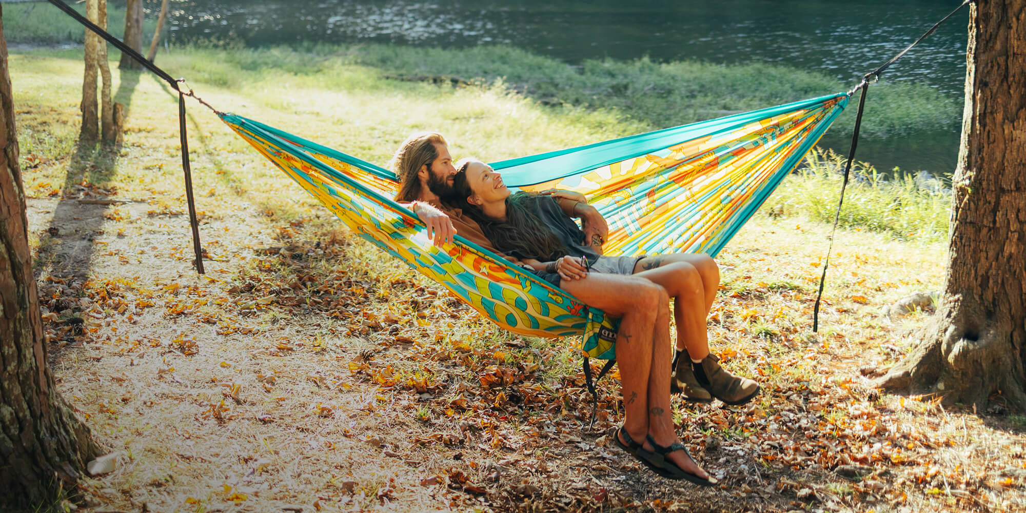 Two people relaxing in a colorful ENO Giving Back hammock by a lake.