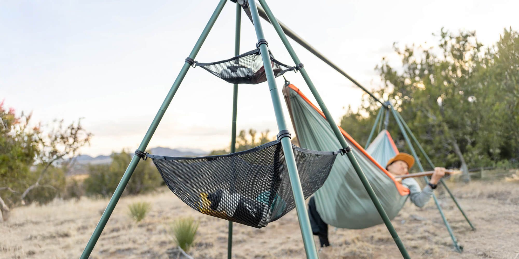 Person lying in an ENO DoubleNest Hammock attached to a Nomad Hammock Stand in a desert landscape with the Nomad Gear Loft setup in the foreground.