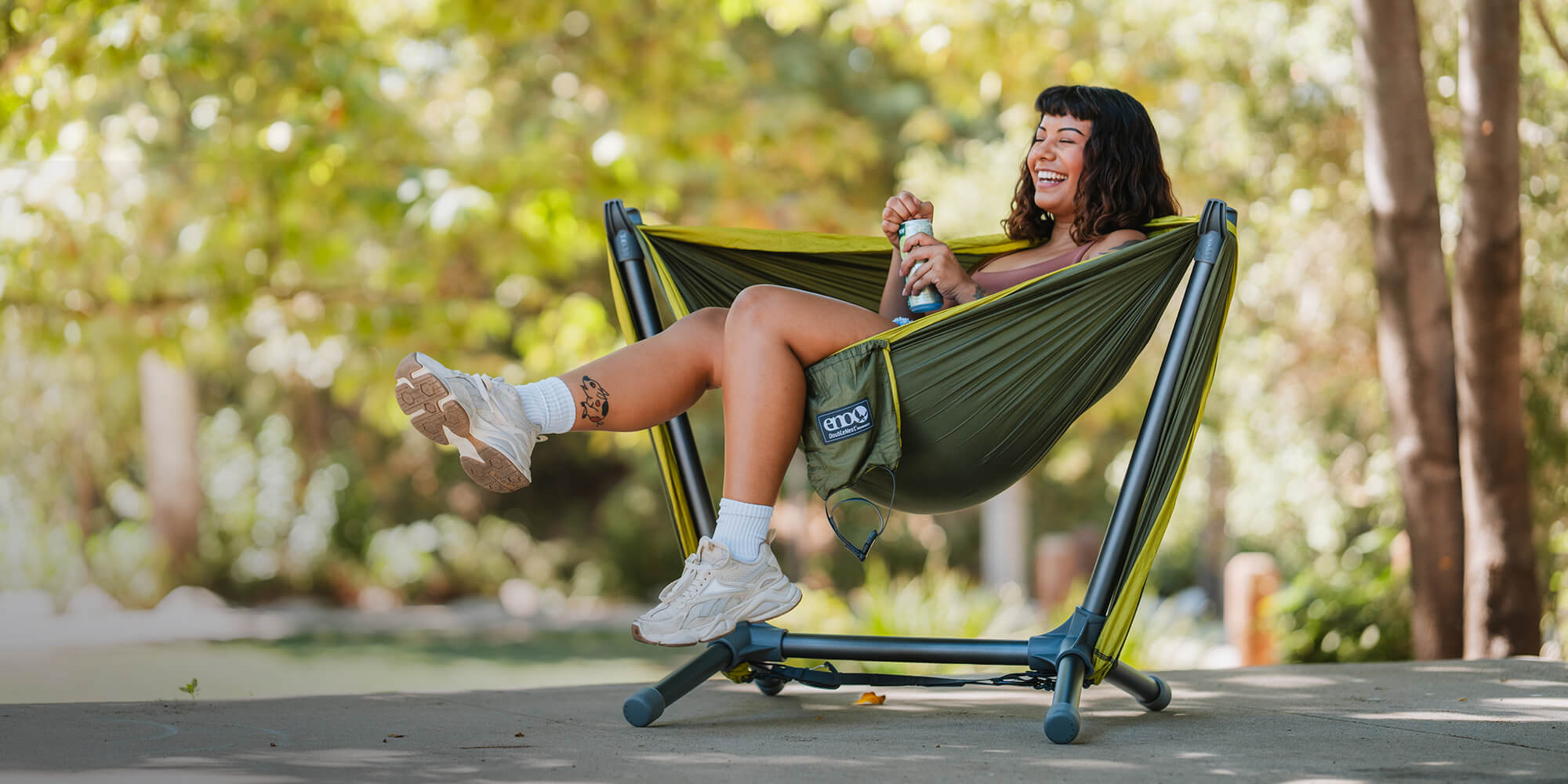 Woman sits smiling in an ENO Parklite Hammock Chair Stand with ENO DoubleNest connected while swinging their legs in from of lush green background.