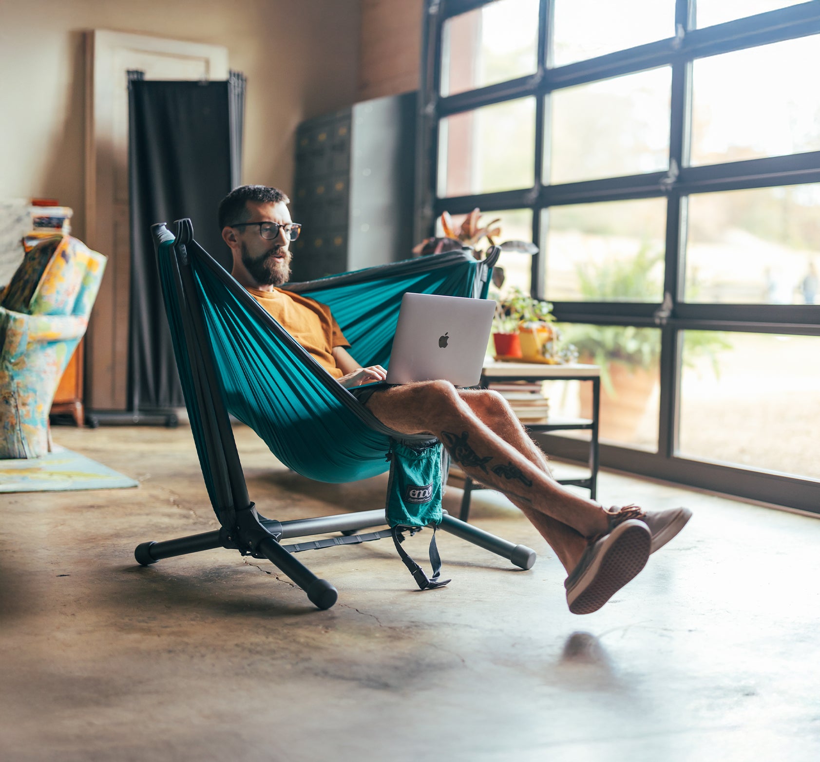 Man using a laptop in a DoubleNest Hammock set up on a ENO Parklite Hammock Chair Stand indoors.