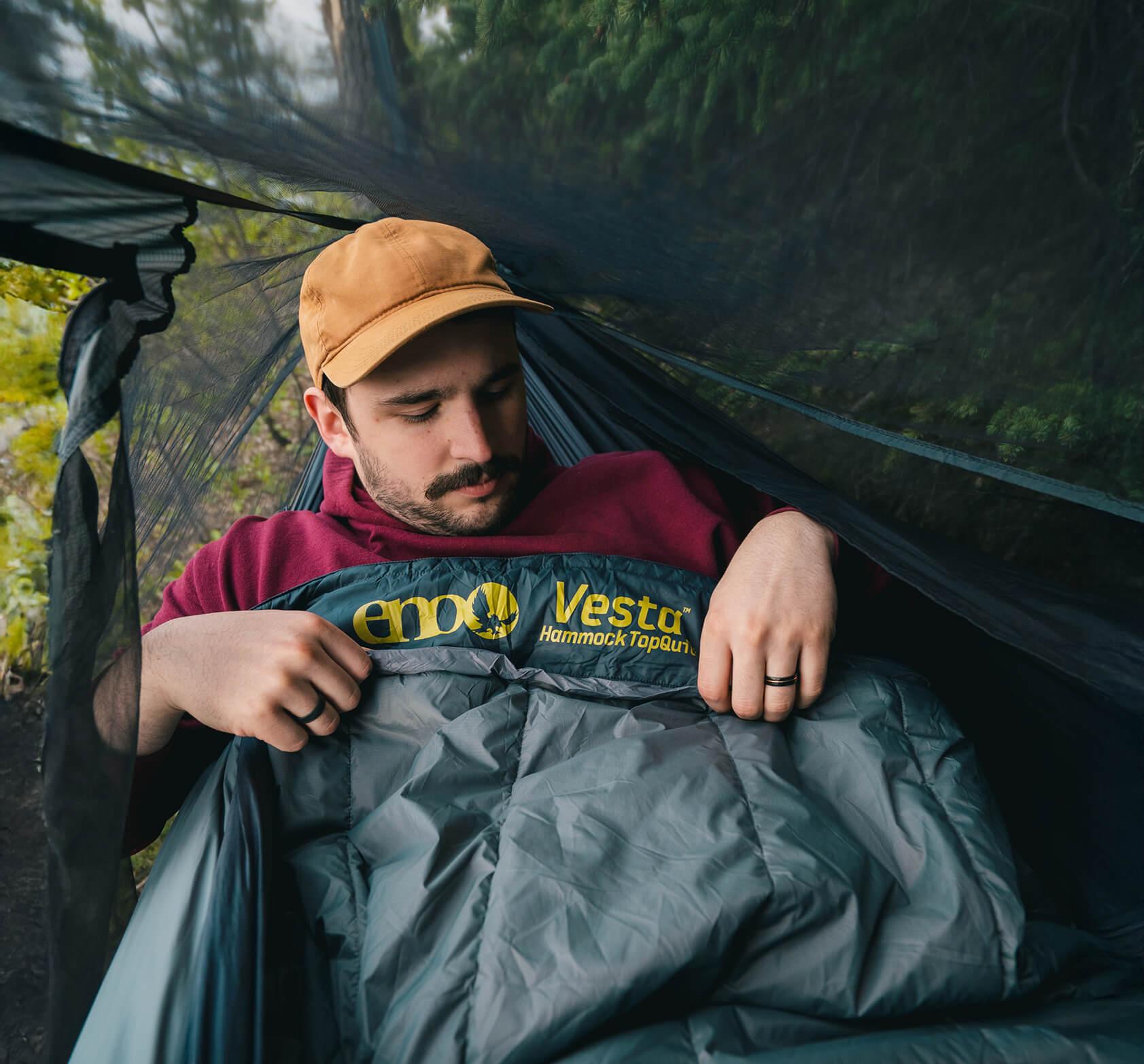 Man sitting in a hammock with a Vesta Hammock TopQuilt, surrounded by greenery and enclosed within an ENO Bug Net.