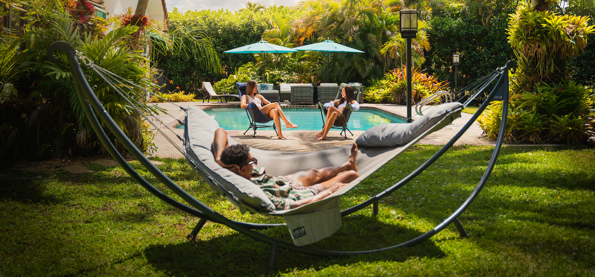 A group of friends relax poolside in their lush, tropical backyard, while two women talk in ENO Lounger DL Chairs while a man lay's back in a ENO SuperNest SL Hammock set up on a SoloPod XL Hammock Stand.