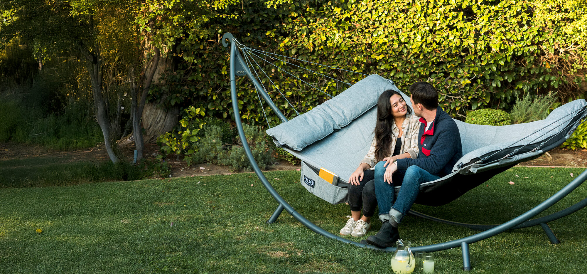 Two people sitting on a SuperNest Hammock set up on a SoloPod XL Hammock Stand in a garden setting.