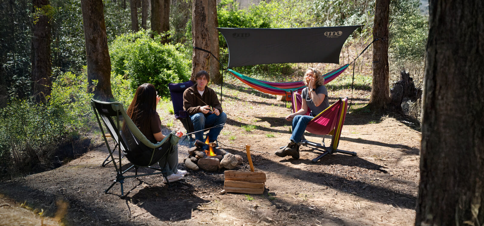 A group of friends sit fireside at their camp site eating s'mores while relaxed in the ENO Lounger DL Mesh Chair, Lounger DL Chair, Parklite Hammock Chair Stand with DoubleNest Hammock, while a DoubleNest Hammock Print under a ProFly Rain Tarp hand g in the wooded background.