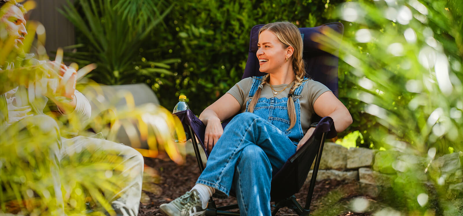 A young women sits smiling surrounded by lush greenery in an ENO Lounger DL Chair with their legs crossed.