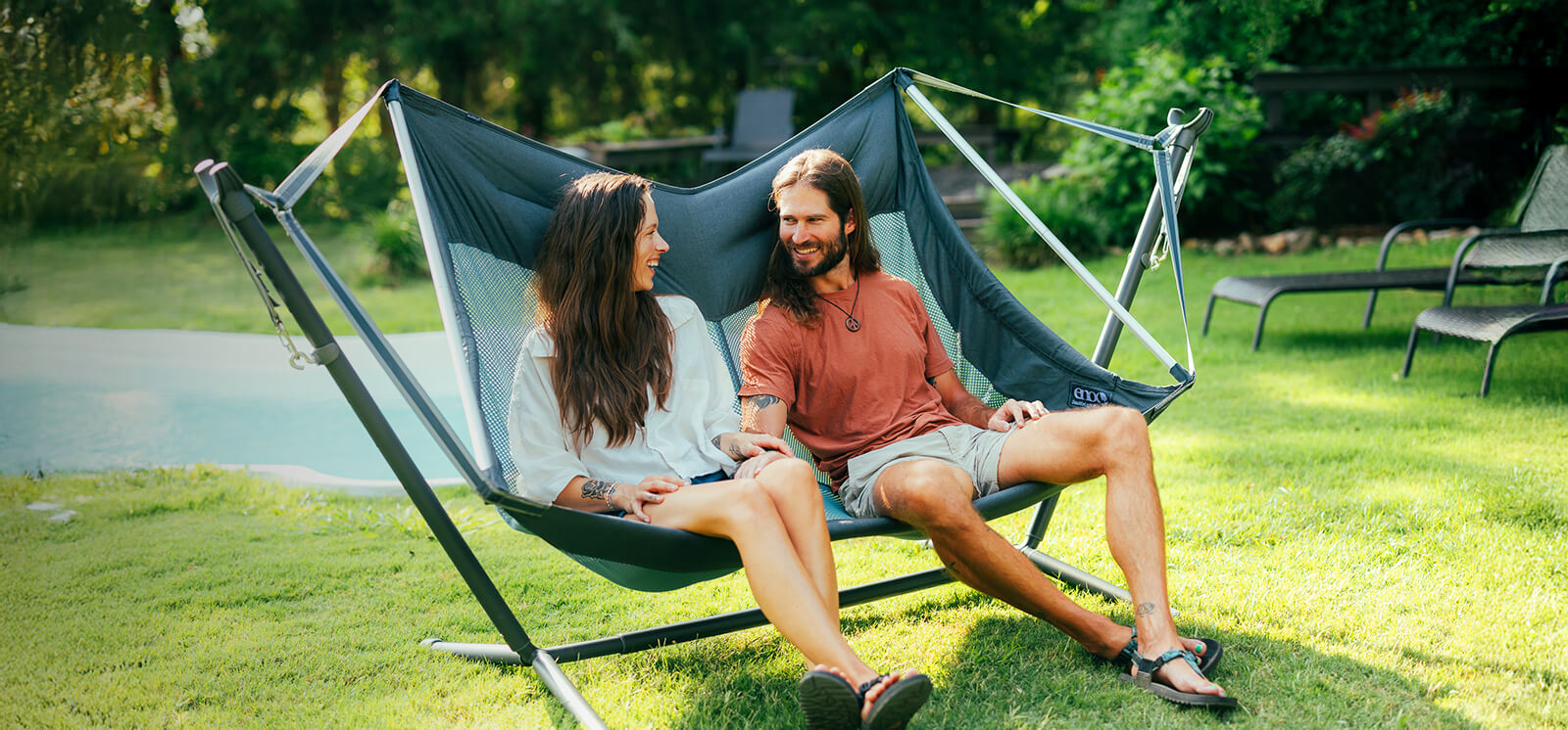 Two people sitting in the DoubleLoft Hammock Chair set up on the Parkway Adjustable Hammock Stand by a pool with a scenic background.