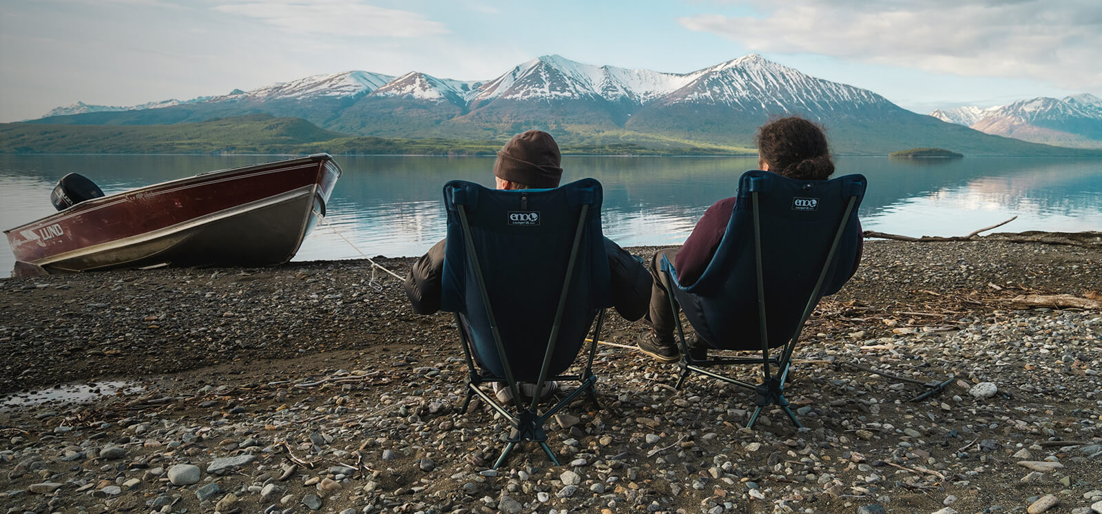 Two people sitting in Lounger DL Chairs by a lake with mountains in the background