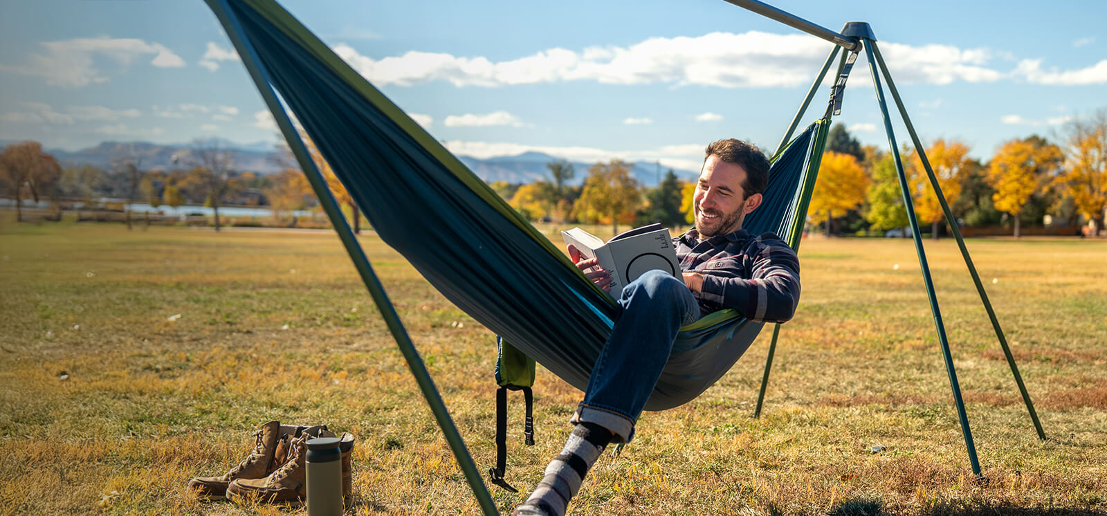 Man reading a book in a Cedar Marine DoubleNest Hammock attached to a Nomad Hammock Stand in a field with mountains in the background.