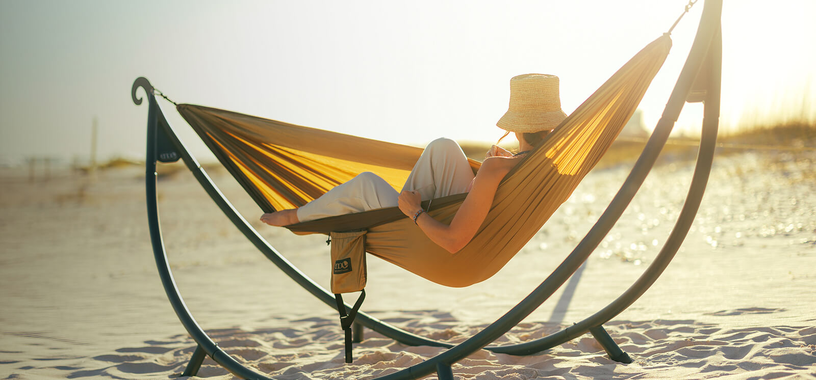 A women rests beachside in a ENO DoubleNest Hammock in Latte Chocolate with the sunsetting in the background.
