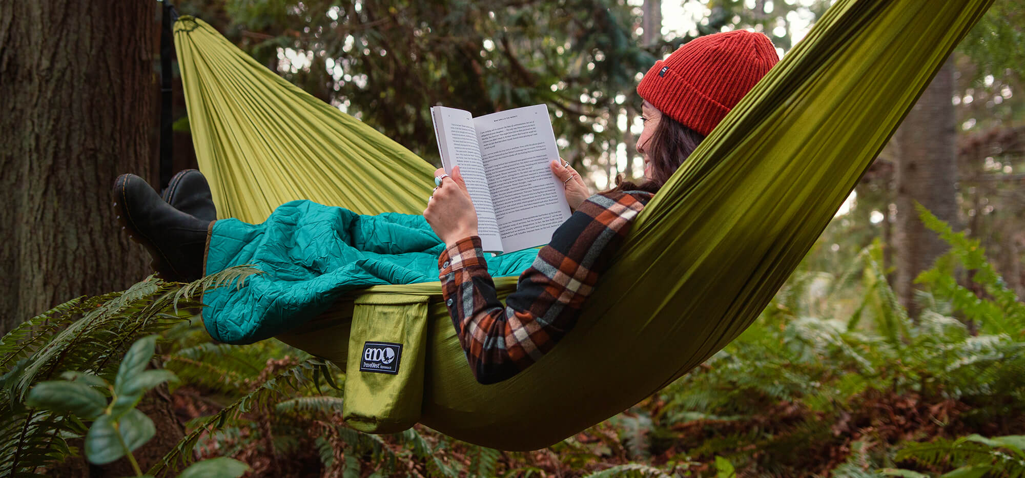 A person smiling while reading a book in a Moss TravelNest Hammock + Straps Combo surrounded by nature.