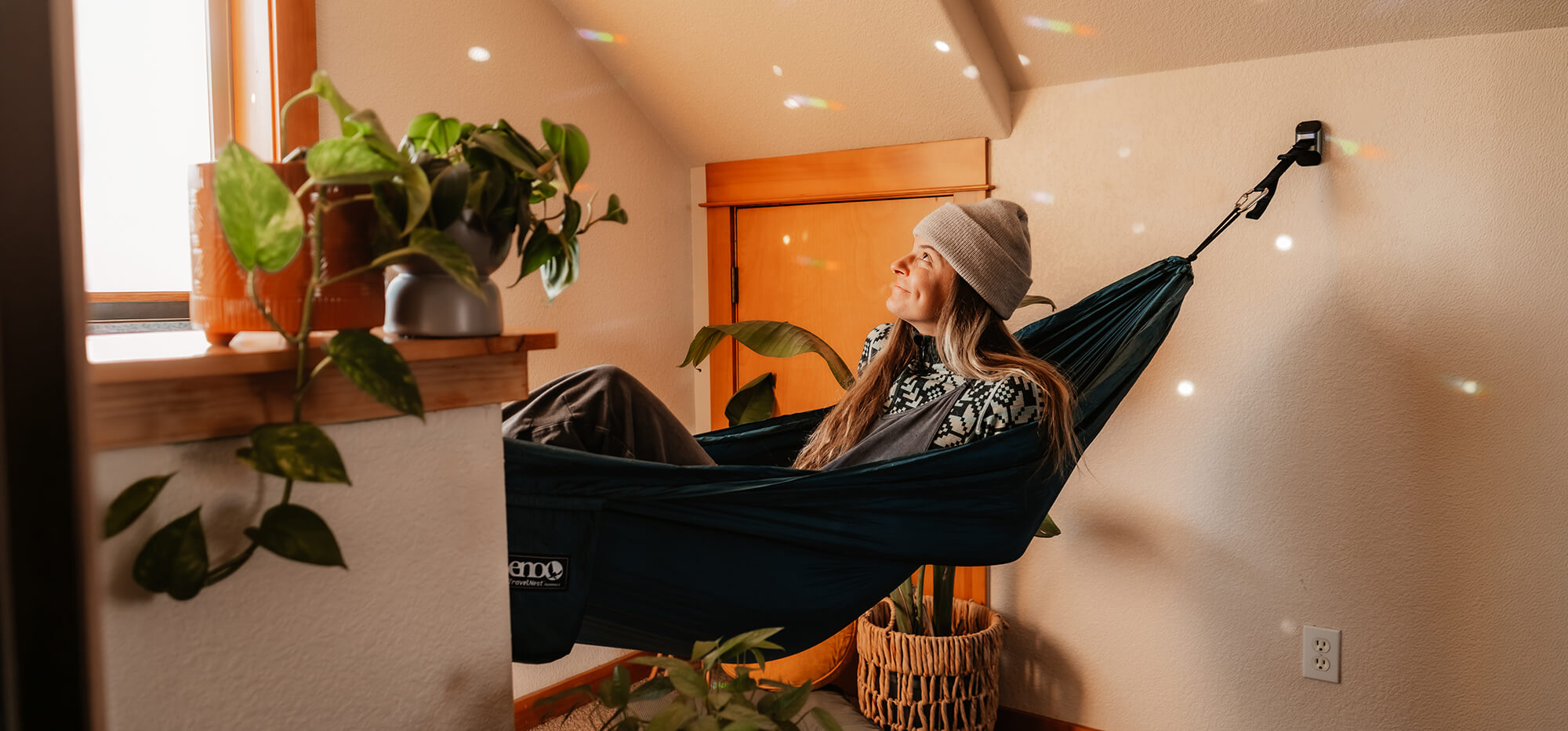 Person relaxing in an ENO TravelNest Hammock attached to the Deluxe Hanging Kit in a cozy room with plants and wooden furniture.
