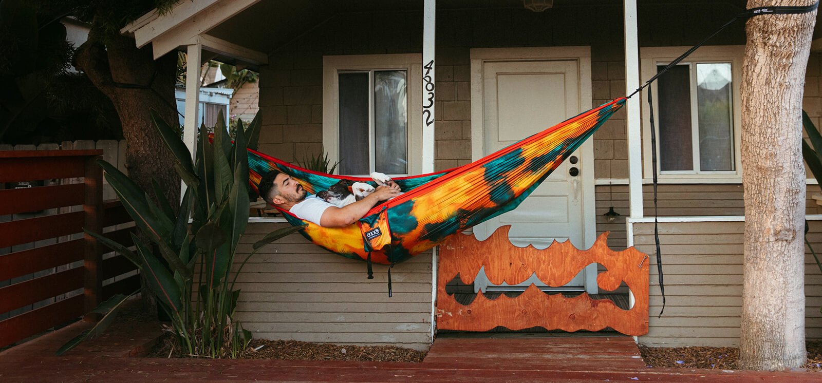 A person rest in an ENO DoubleNest Hammock Print in Tie-Dye on the front porch with his dog laying with them.