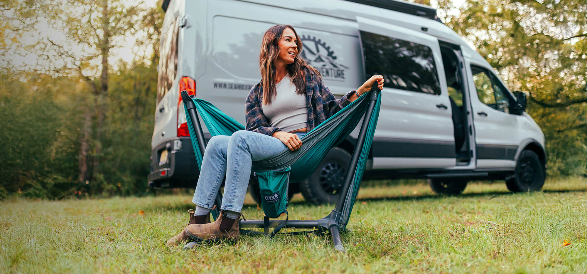 Woman sitting in a Parklite Hammock Chair Stand in front of a camper van in a grassy area.