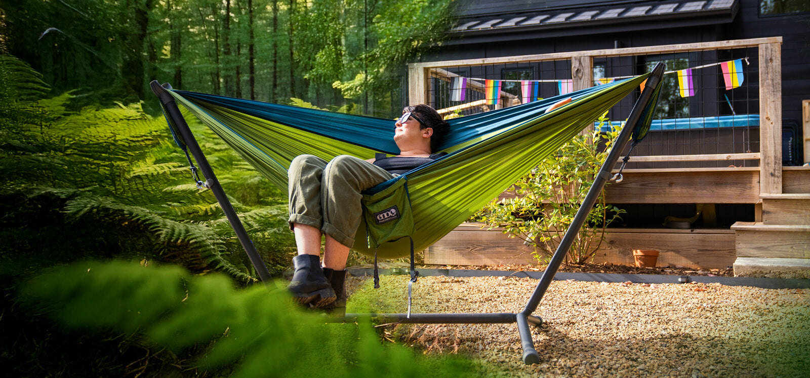 A person sits back relaxing in a ENO DoubleNest Hammock on a Parkway Adjustable Hammock Stand looking into the distance while the background transitions from home to camp.