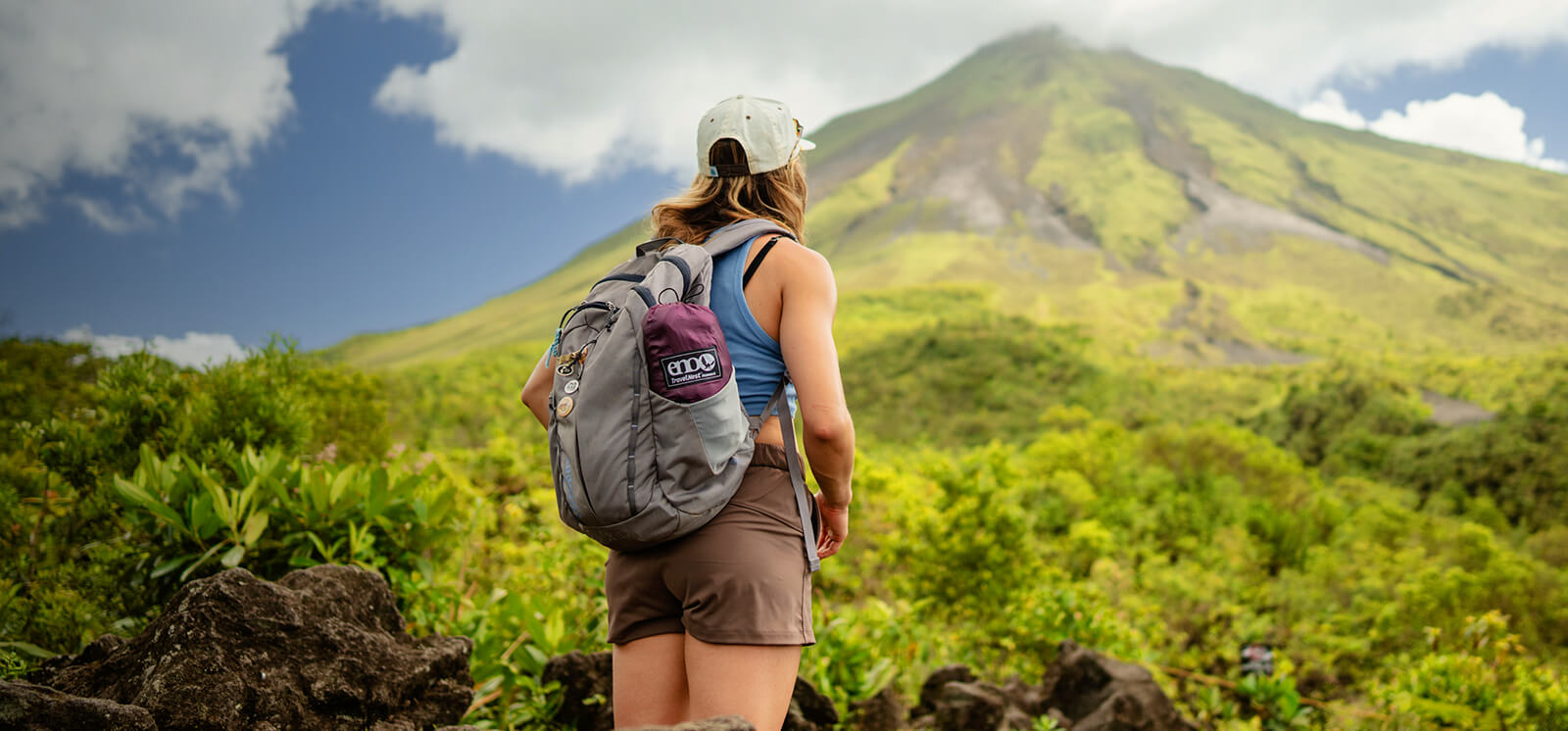 A person looks up at a mountain while hiking, wearing a backpack with an ENO TravelNest Hammock + Strap Combo in side pocket.