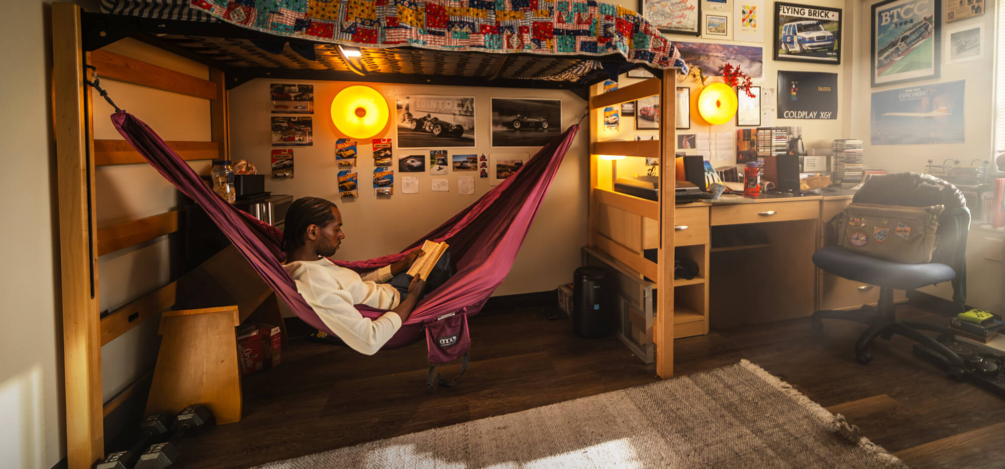 A person relaxes in a DoubleNest Hammock suspended under a wooden loft bed in a cozy, decorated dorm room setting. The room features a desk, chair, and warm lighting from hanging lamps.