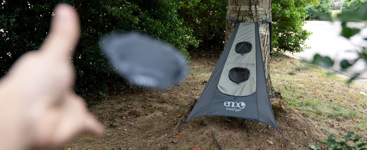 A person playing the new TrailFlyer Outdoor Game in Charcoal-Sage suspended from a tree outdoors, with a blurred hand and disc visible in the foreground.
