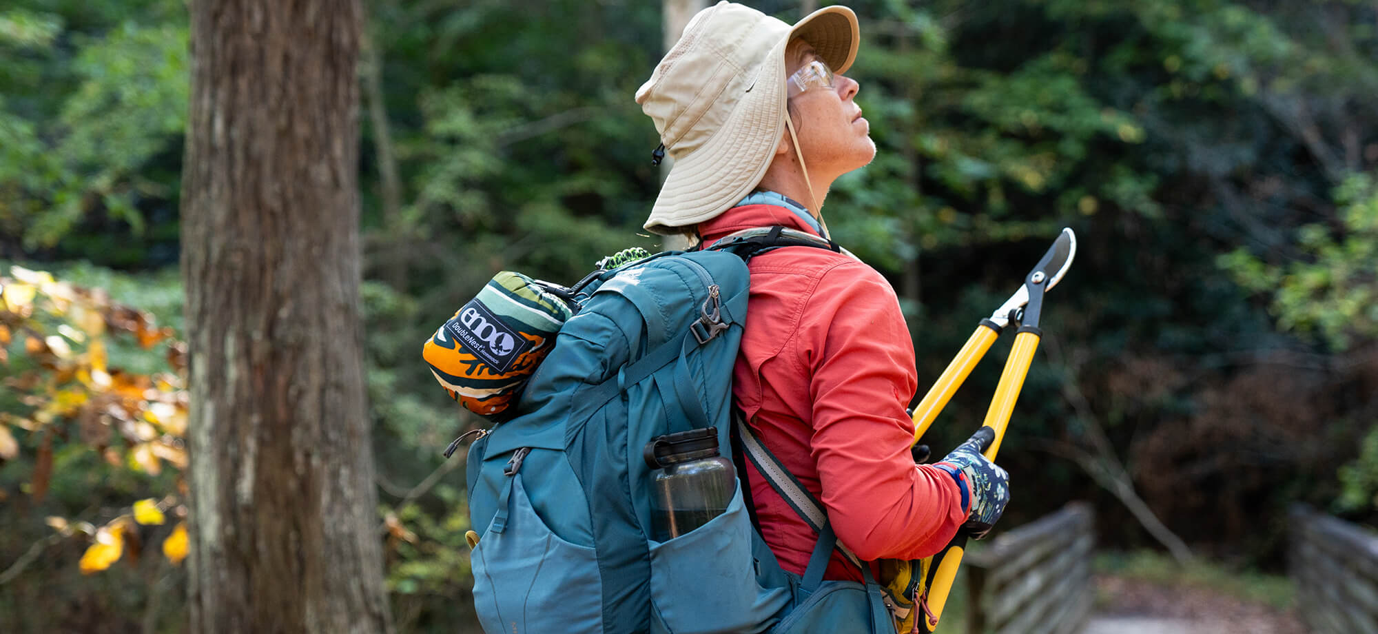A person hiking with a backpack and trimming shears in a forest with a packed ENO Giving Back Hammock attached to the bag