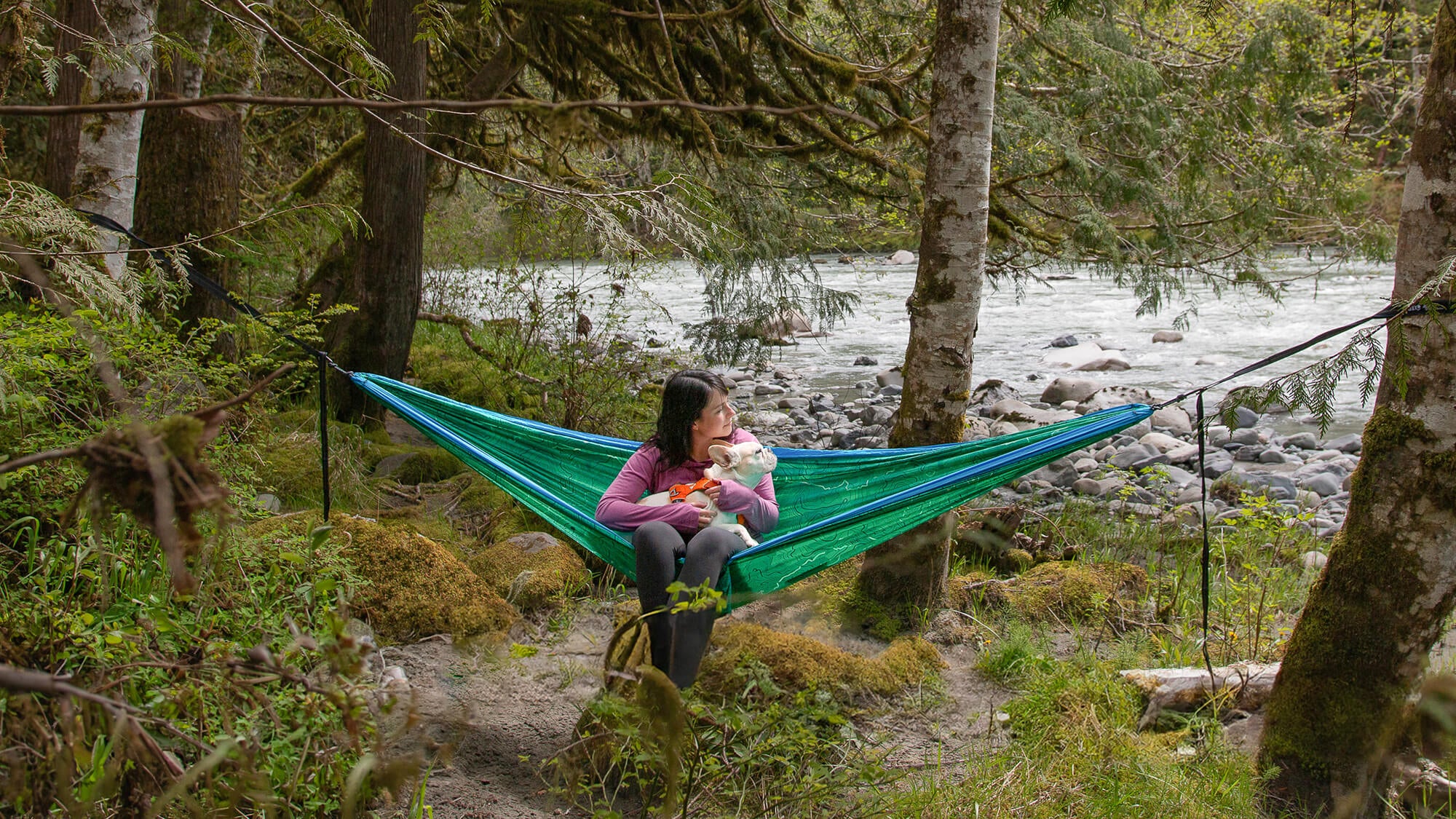 Woman sits with white dog in lap in the ENO DoubleNest PCT Giving Back Hammock while looking out onto river in the background.