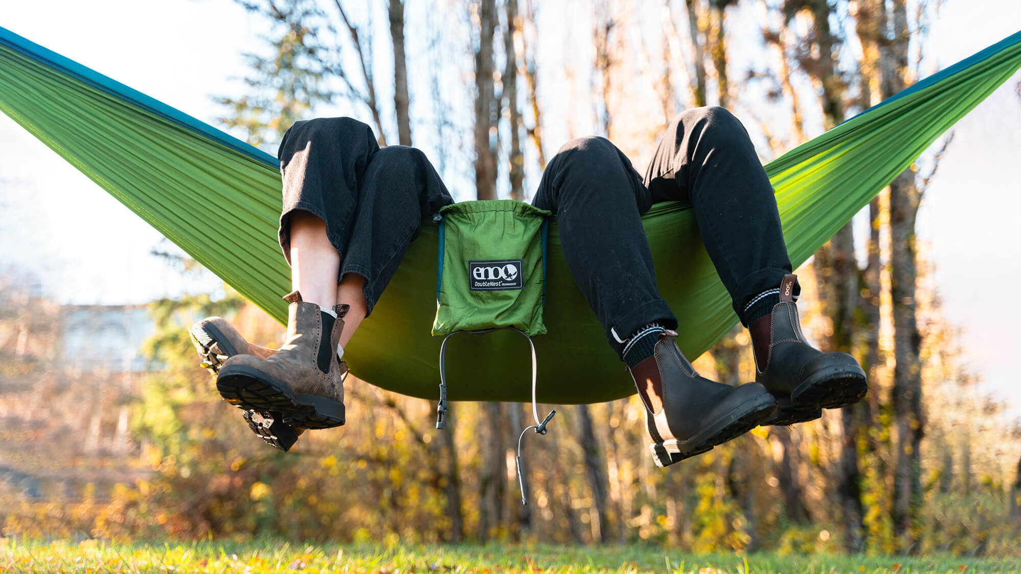 Two people sit in the ENO DoubleNest Hammock in Cedar Marine colorway with their legs hanging out.