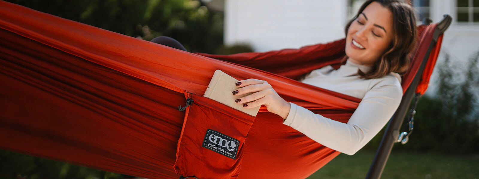 Women rests in an ENO DoubleNest Hammock set up on a Parkway Hammock Stand in backyard while they grab a book from the hammock stuff sack pocket.