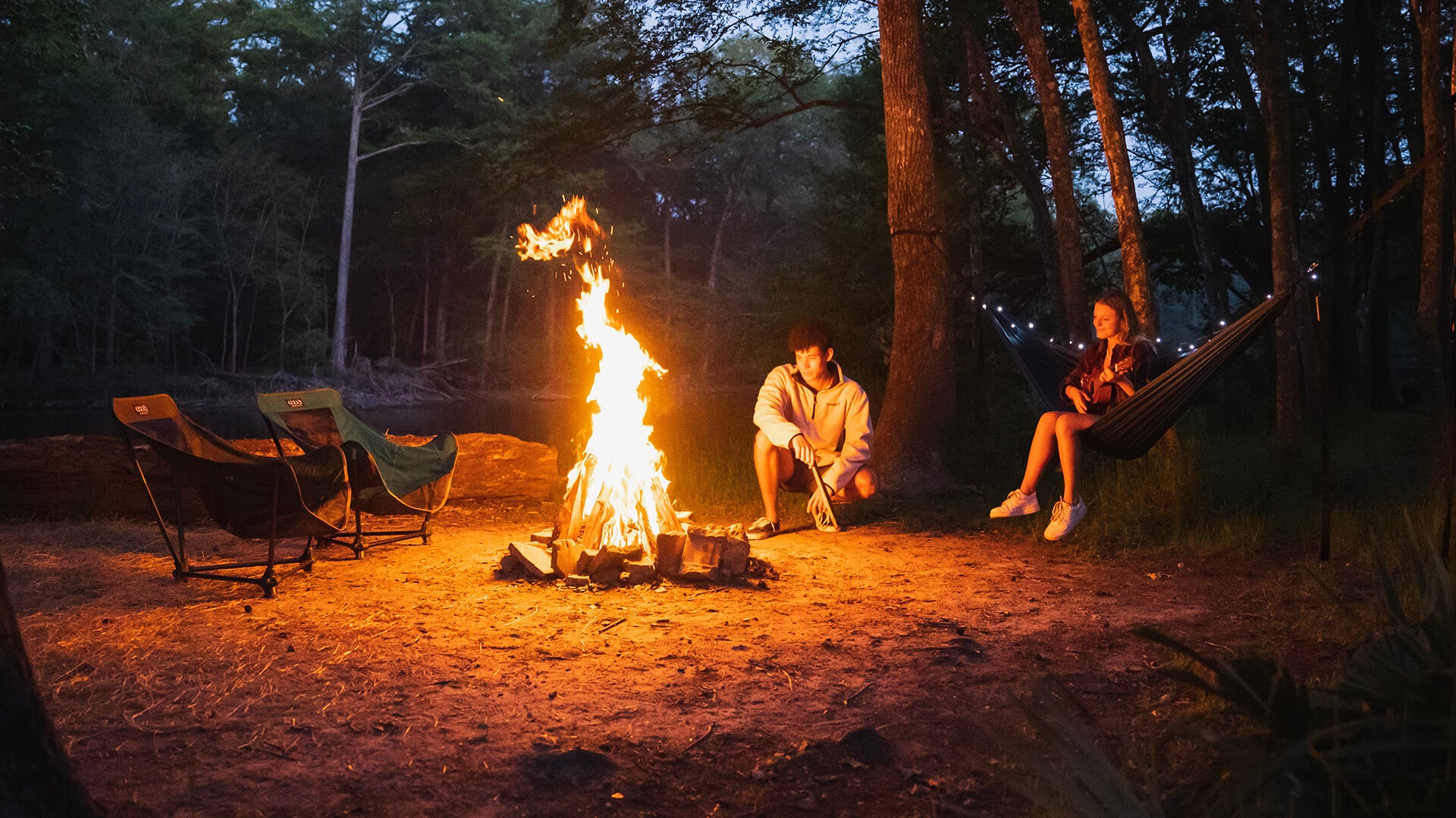 A young couple relaxes beside a large campfire at a campsite next to a river with ENO chairs and an ENO hammock.