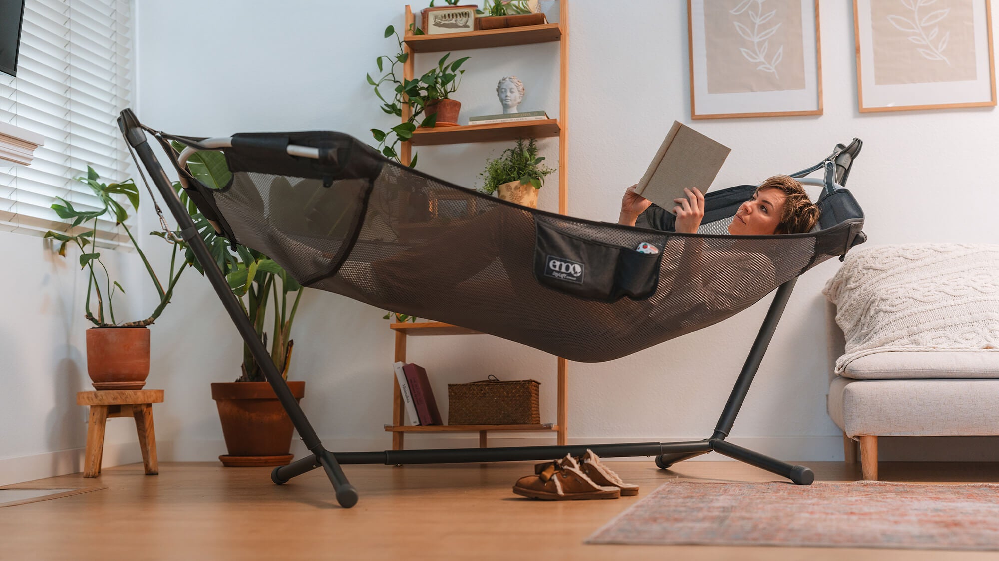 Woman laying down relaxing in an ENO DayLoft Hammock hanging in a Parklite Adjustable Hammock Stand while reading a book in the living room.