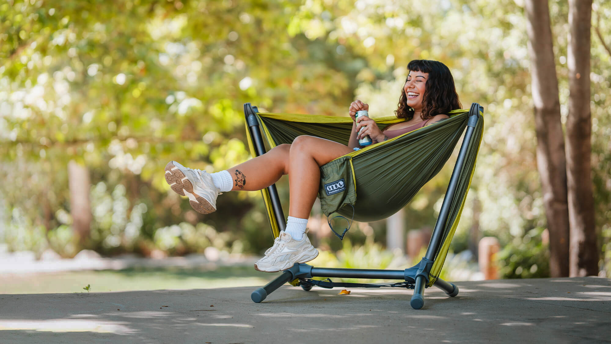 Woman sits back, kicking a leg up with beverage in hand, in the ENO Parklite Hammock Chair Stand with DoubleNest Hammock in front of lush green backdrop. 