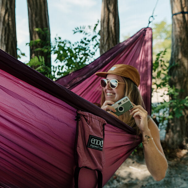 A woman in hat and sunglasses looks out of their ENO portable hammock while holding a camera.