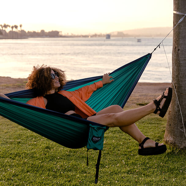 A woman swings her legs out of an ENO portable hammock in park setting with waterway and shore in the background. 